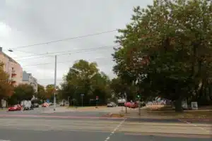Straßenbild, Kreuzung von drei Straßen, großer Baum am rechten Bildrand, links ein Wohngebäude
