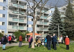 Menschen stehen auf einer Wiese, dahinter ein orangefarbener Pavillion, ein Cafe auf Rädern sowie die soeben eingewihte Erzählbank