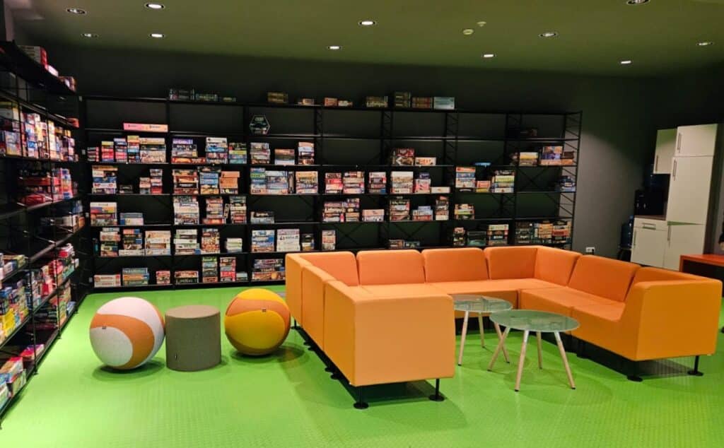 Ein Raum mit grünem Fußboden. In der Mitte ein großes orangefarbiges Sofa in U-Form aufgestellt. Links neben dem Sofa befinden sich zwei Sitzbälle, ebenfalls mit orangen Farbtönen. An der linken Wandseite sowie der dem Betrachter gegenüberliegenden Wand stehen Raumhohe Wandregale befüllt mit unterschiedlichen Brettspielsammlungen.