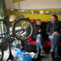 Bild mit zwei Personen, die sich bei einem Kaffee unterhalten, während ein Kinderfahrrad repariert wird.