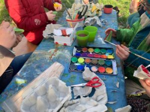 Mit Farbe, Eierkartons, Pinseln und Scheren belegter Tisch auf einer Wiese, an dem Kinder sitzen