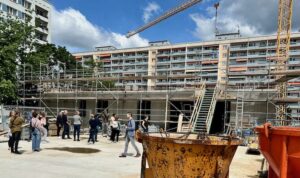 Baugeräte und Kran vor dem flachen Rohbau, dahinter ein 10-geschossiger Plattenbau aus den 1970er Jahren