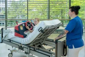 Schwester Katrin schiebt Greta in ihrem Patientenbett über den Krankenhausflur. Das mobile Cockpit lenkt Greta dabei ab und beschäftigt sie.