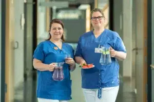 Zwei Frauen in Berufskleidung halten Wasserkrüge sowie Obstteller in ihren Händen.