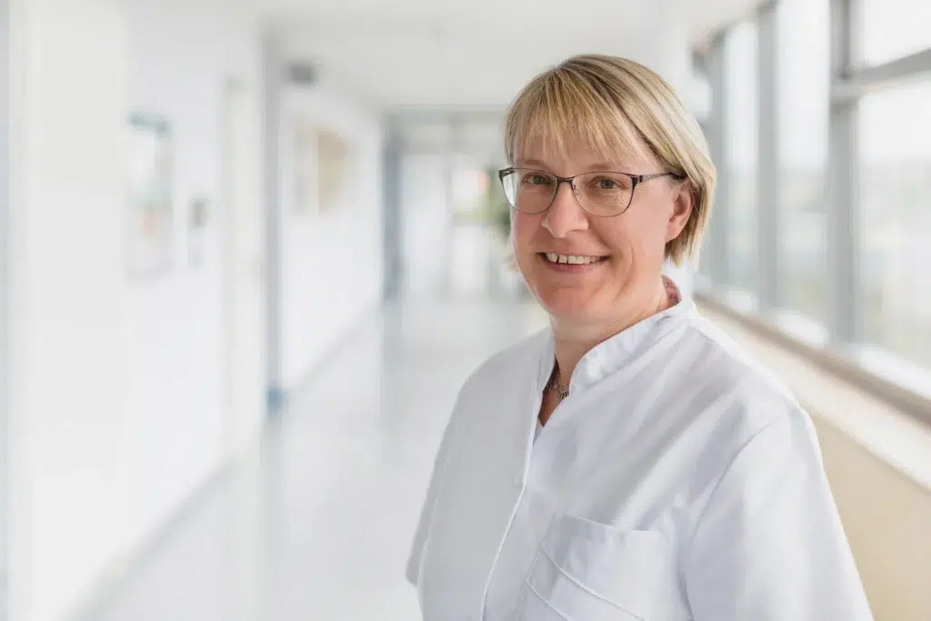Frau mittleren Alters, Brille, blonde Haare, freundlich und mit weißem Arztkittel bekleidet.